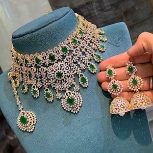 Könnte beinhalten: Ein goldfarbenes Halsketten- und Ohrring-Set mit klaren und smaragdgrünen Edelsteinen. Die Halskette hat ein florales Design mit tropfenförmigen Smaragden. Die Ohrringe haben ein ähnliches Design mit einem glockenförmigen Boden und einer Perle.