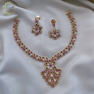 Può includere: Un set di collana e orecchini in oro con un design floreale. La collana presenta un grande ciondolo con un gruppo di pietre rosa e bianche. Gli orecchini sono più piccoli e hanno un'unica pietra rosa al centro.