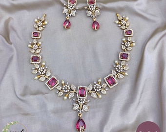 May include: A gold necklace with a pink stone pendant and matching earrings. The necklace features a floral design with white stones and pink rectangular stones. The earrings have a similar design with a pink stone drop.