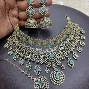 May include: A gold-toned jewelry set featuring a necklace, earrings, and a headpiece. The set is adorned with clear and emerald-green gemstones. The earrings have a chandelier design, and the necklace has a floral pattern.
