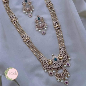 Puede incluir: Un collar de oro con tres hileras de cuentas y un gran colgante con un diseño de pavo real con piedras verdes y blancas. El colgante está adornado con pequeñas gotas de perlas y piedras rojas. También se incluyen pendientes a juego con un diseño de pavo real y piedras verdes y blancas.