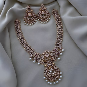 May include: A gold necklace with a large pendant featuring intricate floral designs and white stones. The necklace is adorned with smaller white stones and pearls. Two matching earrings with similar designs are also included.
