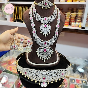 Puede incluir: Un conjunto de joyas de novia indias tradicionales que incluye un collar blanco y plateado con piedras rosas, un collar a juego, pendientes y un cinturón de cintura. El conjunto se muestra en un soporte negro.
