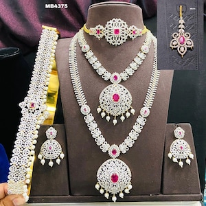 May include: A bridal jewelry set with a gold base and white stones. The set includes a necklace, earrings, a maang tikka, and a waist belt. The necklace has three strands with a large pendant in the center. The earrings are teardrop shaped with a single large stone. The maang tikka is a traditional Indian headpiece with a single large stone. The waist belt is a long, thin chain with a single large stone in the center.