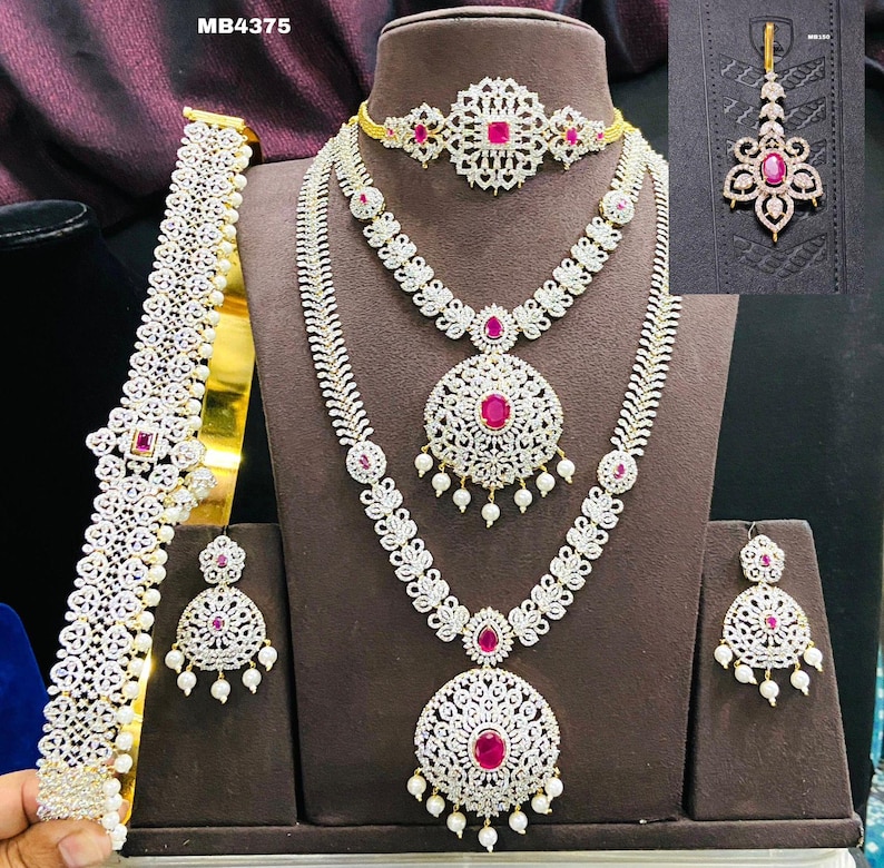 May include: A bridal jewelry set with a gold base and white stones. The set includes a necklace, earrings, a maang tikka, and a waist belt. The necklace has three strands with a large pendant in the center. The earrings are teardrop shaped with a single large stone. The maang tikka is a traditional Indian headpiece with a single large stone. The waist belt is a long, thin chain with a single large stone in the center.