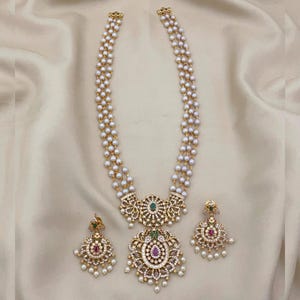 Op de afbeelding: Een gouden ketting- en oorbellenset met meerdere strengen kleine, witte parels. De ketting heeft een grote, sierlijke hanger met een centrale groene steen en omringende heldere stenen. De oorbellen weerspiegelen het hangerontwerp.