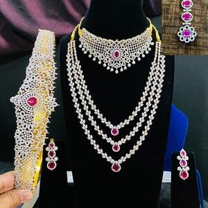 May include: A gold and white bridal jewelry set with pink stones. The set includes a necklace, earrings, a headpiece, and a choker.