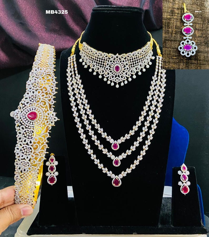 May include: A gold and white bridal jewelry set with pink stones. The set includes a necklace, earrings, a headpiece, and a choker.