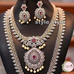 May include: A two-piece bridal necklace set with a long and short chain. The necklace is made of gold and features a large, round pendant with a red stone in the centre. The pendant is surrounded by smaller stones. The necklace is also adorned with pearls.