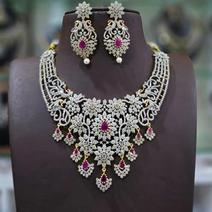 Könnte beinhalten: Ein silberfarbenes Halsketten- und Ohrringset mit komplizierten, von Blumen und Pfauen inspirierten Filigranmustern. Der Schmuck ist mit zahlreichen klaren weißen Steinen besetzt und mit mehreren tropfenförmigen rosa Steinen akzentuiert.