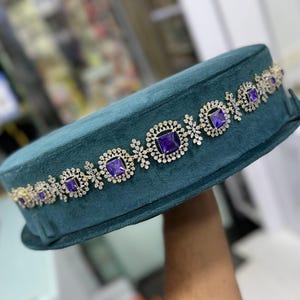 Könnte beinhalten: Ein teal-farbener Samthut mit einem dekorativen Band aus quadratischen, violetten Edelsteinen und klaren Kristallen. Das Band ist in einem goldfarbenen Metall gefasst. Der Hut hat eine abgerundete, rechteckige Form.