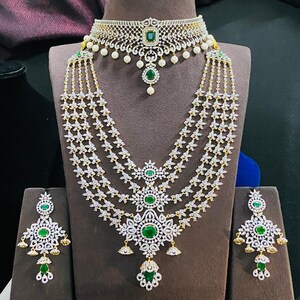 May include: A three-strand diamond necklace set with green emerald accents. The set includes a choker necklace, earrings, and a pendant. The necklace has a floral design and the earrings are teardrop shaped.