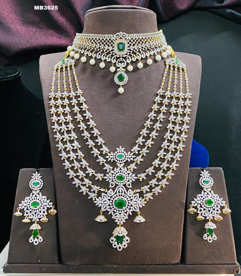 May include: A three-strand diamond necklace set with green emerald accents. The set includes a choker necklace, earrings, and a pendant. The necklace has a floral design and the earrings are teardrop shaped.