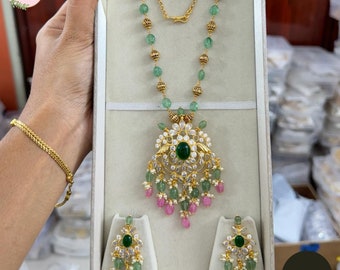 May include: A gold-toned necklace and matching earrings set. The necklace features green and gold beads, with a large pendant adorned with a central green stone, white accents, and pink and green dangles. The earrings mirror the pendant design. The set is displayed in a gray box.