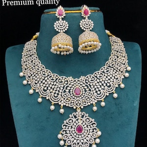 Puede incluir: Un deslumbrante conjunto de joyas con collar y pendientes a juego. El collar y los pendientes están adornados con cristales transparentes, perlas de imitación y piedras preciosas de color rubí. El texto "NJ 384 Gj finish Premium quality" es visible.