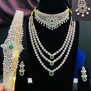 May include: A traditional Indian bridal jewelry set featuring a gold necklace with three strands of white stones and green accents, a matching choker, a headband, and earrings. The set also includes a traditional nose ring with white stones and a green accent.