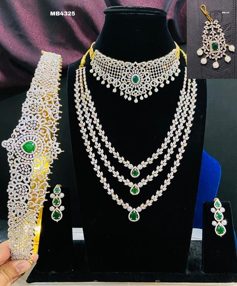 May include: A traditional Indian bridal jewelry set featuring a gold necklace with three strands of white stones and green accents, a matching choker, a headband, and earrings. The set also includes a traditional nose ring with white stones and a green accent.