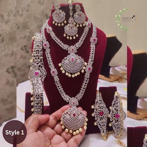 May include: A traditional Indian bridal jewelry set featuring a long, layered necklace with a large pendant, a matching pair of earrings, a maang tikka, a waist chain, and a pair of bangles. The jewelry is made of silver and is adorned with white stones and pink accents. The set is displayed on a red velvet background.  Style 1.