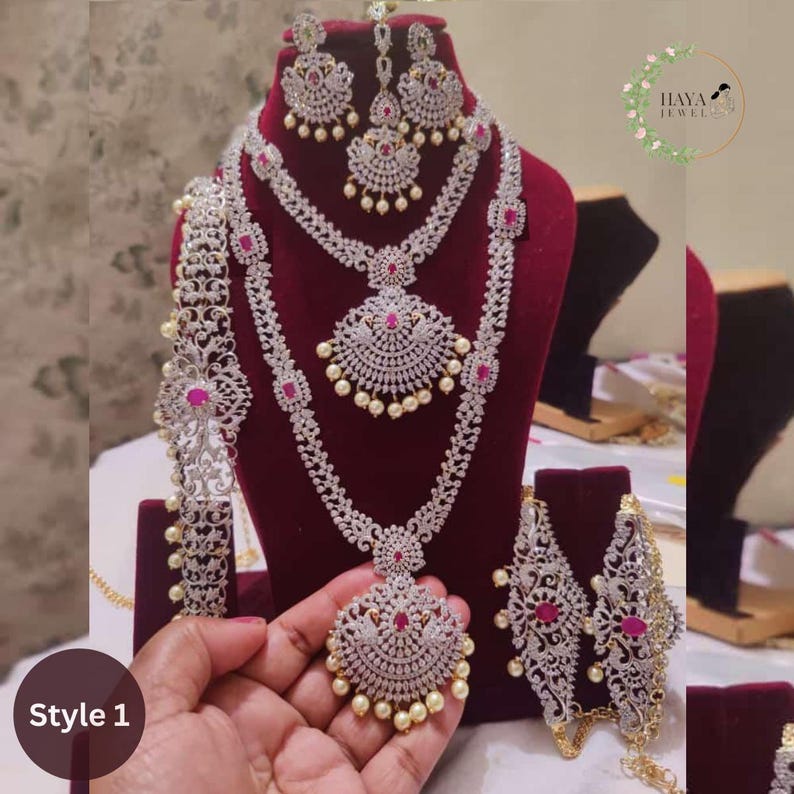 May include: A traditional Indian bridal jewelry set featuring a long, layered necklace with a large pendant, a matching pair of earrings, a maang tikka, a waist chain, and a pair of bangles. The jewelry is made of silver and is adorned with white stones and pink accents. The set is displayed on a red velvet background.  Style 1.