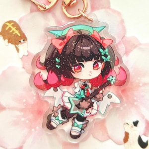 以下が含まれることがあります： ギターを演奏するちびアニメキャラクターが描かれた、ローズゴールドの留め金が付いた透明なアクリルキーホルダー。キャラクターは赤いアクセントのある黒髪で、白とティールの衣装を着ています。キーホルダーはピンクの花びらの背景に配置されています。