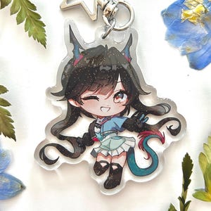 Può includere: Un portachiavi in acrilico trasparente con un personaggio in stile anime con capelli neri, dettagli blu e rossi e una coda turchese. Il portachiavi ha una parte superiore a forma di stella argentata e una chiusura argentata. Lo sfondo include fiori secchi e vegetazione.