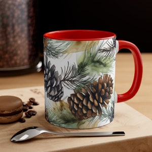 May include: A red and white ceramic mug with a watercolor design of pine branches and pine cones. The mug has a red rim and handle.