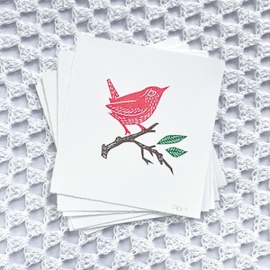 May include: A stack of red and green illustrated cards featuring a red bird perched on a branch with green leaves. The cards are on a white crocheted surface.
