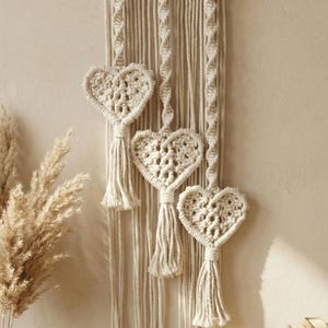 Puede incluir: Colgante de pared de macramé color crema con tres corazones calados y borlas. La pieza está suspendida de un listón de madera, mostrando un estilo bohemio. El fondo es beige neutro.