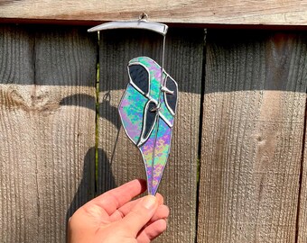 Puede incluir: Un atrapasoles de vidriera de la Parca con una guadaña y colores iridiscentes de púrpura, verde y azul. La pieza decorativa está suspendida por un alambre de metal plateado.