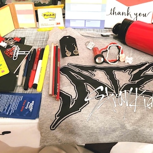 May include: A gray t-shirt with black lettering that says "Stray Kids" and a red water bottle. The t-shirt also has a black and white graphic design. There are various school supplies on the table, including a ruler, pencils, pens, and a small notepad.