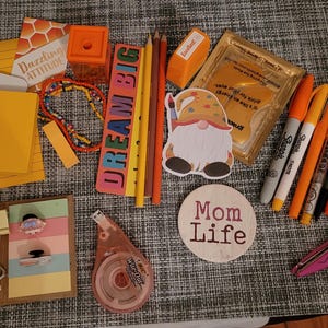 May include: A flat lay of various craft supplies, including colorful pencils, pens, a gnome sticker, a ruler, a tape dispenser, a pair of scissors, and a wooden sign that says "Mom Life".