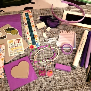 May include: A collection of purple and pink stationery supplies, including stickers, pens, a ruler, a notepad, and a headband. The items are arranged on a textured surface with a brown background.