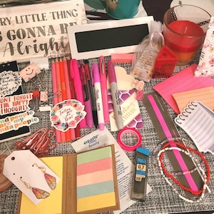 May include: A collection of colorful stationery supplies, including pens, pencils, stickers, and notepads. The items are arranged on a gray textured surface. The text "EVERY LITTLE THING IS GONNA BE Alright" is visible on a wooden sign.