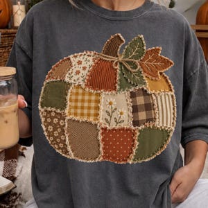 May include: A charcoal gray t-shirt featuring a large, central pumpkin graphic composed of various fabric patches in shades of orange, brown, green, and cream. The pumpkin design includes floral, gingham, striped, and polka dot patterns.
