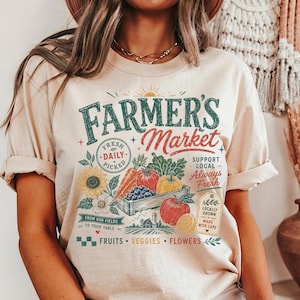 May include: A beige t-shirt featuring a graphic print with the text "FARMER'S Market" in green and red. The design includes illustrations of a wooden crate filled with carrots, tomatoes, blueberries, and peppers, alongside a sunflower and text.