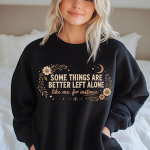 Pode incluir: Uma camisola preta de gola redonda com um design gráfico que inclui o texto "SOME THINGS ARE BETTER LEFT ALONE like me, for instance." O design é rodeado por ilustrações florais, pequenas estrelas e uma lua crescente em tons de bege e castanho.
