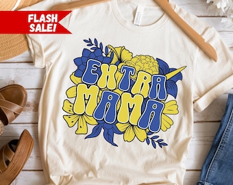 May include: A cream colored t-shirt with a floral design and the text "EXTRA MAMA" in blue and yellow letters. The shirt is hanging on a wooden hanger.