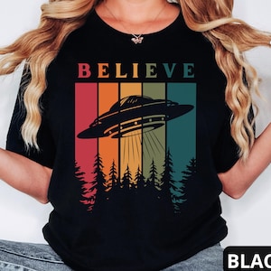 Puede incluir: Camiseta negra con un gráfico retro de un OVNI volando sobre un bosque con la palabra "BELIEVE" encima.
