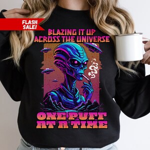 May include: Black sweatshirt with a graphic of an alien smoking a cigarette. The text reads "Blazing it up across the universe one puff at a time".