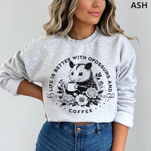 May include: A gray sweatshirt with a black graphic print of an opossum holding a cup of coffee. The graphic also includes the text "Life is better with opossums and coffee".
