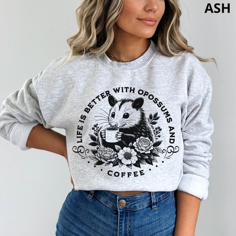May include: A gray sweatshirt with a black graphic print of an opossum holding a cup of coffee. The graphic also includes the text "Life is better with opossums and coffee".