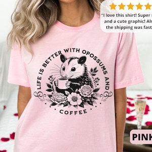 May include: Pink t-shirt with a black and white graphic of an opossum holding a cup of coffee. The text on the shirt reads "Life is better with opossums and coffee."