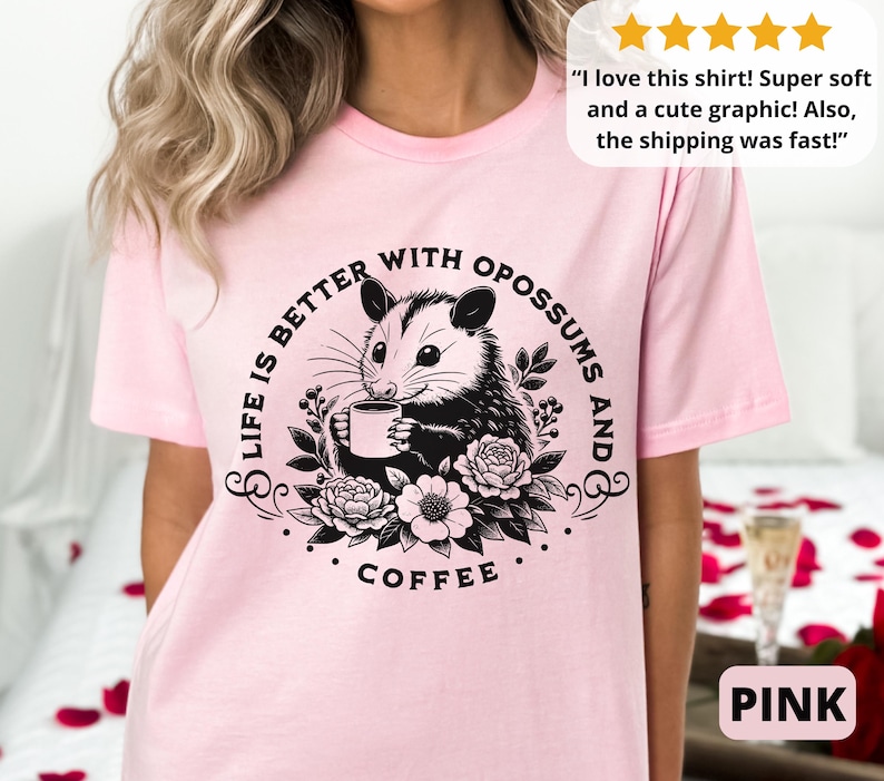 May include: Pink t-shirt with a black and white graphic of an opossum holding a cup of coffee. The text on the shirt reads "Life is better with opossums and coffee."