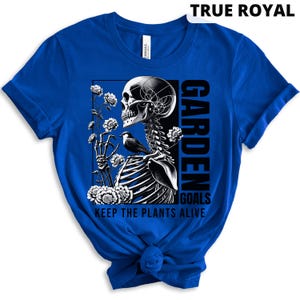 May include: A royal blue t-shirt with a black and white graphic of a skeleton holding flowers and a bird. The text "Garden Goals Keep the Plants Alive" is printed on the shirt.