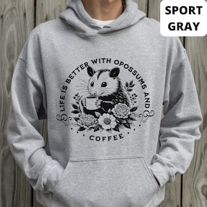 May include: A gray hooded sweatshirt with a black and white graphic of an opossum holding a cup of coffee. The graphic says "Life is better with opossums and coffee".