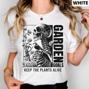 May include: White t-shirt with a black and white graphic of a skeleton holding flowers and a bird. The text "GARDEN GOALS KEEP THE PLANTS ALIVE" is printed on the shirt.