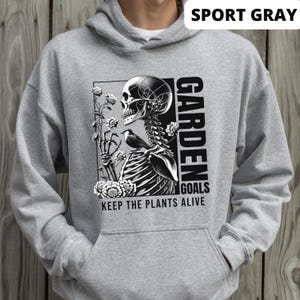 May include: A gray hooded sweatshirt with a black and white graphic print of a skeleton holding flowers and a bird. The text "GARDEN GOALS KEEP THE PLANTS ALIVE" is printed below the graphic. The sweatshirt is labeled "SPORT GRAY".