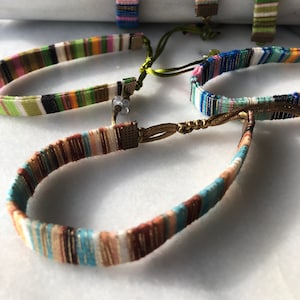 May include: A set of five colourful woven bracelets with gold accents and adjustable closures. The bracelets are made of thin, colourful threads and have a bohemian style.