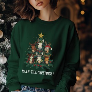 Puede incluir: Una sudadera verde oscuro con un diseño de árbol de Navidad hecho con caras de burro. Los burros están decorados con elementos festivos, y una estrella dorada corona el árbol. El texto "Mule-Tide Greetings!" está impreso debajo.