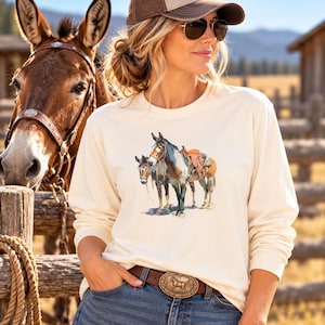 May include: A cream-colored long-sleeve shirt featuring a graphic illustration of two mules, one equipped with a saddle. The shirt is paired with blue denim jeans and a brown leather belt with a large, ornate, oval-shaped metal belt buckle.